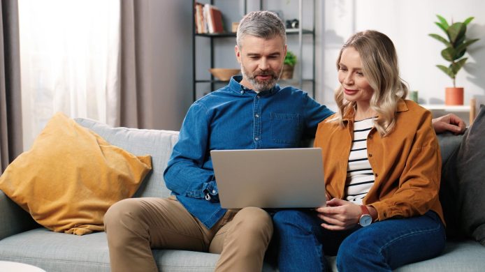 Caucasian middle-aged happy married couple bearded man and beautiful woman sitting on couch in good mood typing on laptop browsing online searching internet at home in cozy room, leisure concept