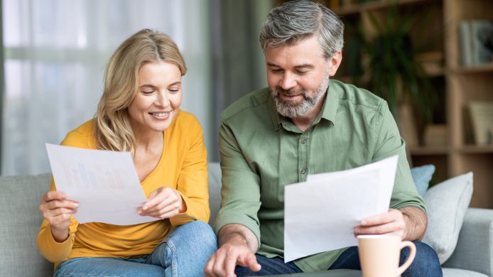 Happy middle aged spouses checking financial documentation and calculating spents while planning family budget together at home, smiling mature spouses reading papers and using calculator