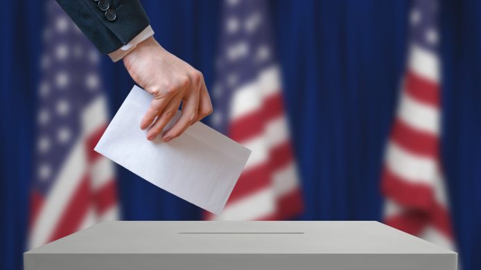 Election in United States of America. Voter holds envelope in hand above vote ballot. USA flags in background. Democracy concept.